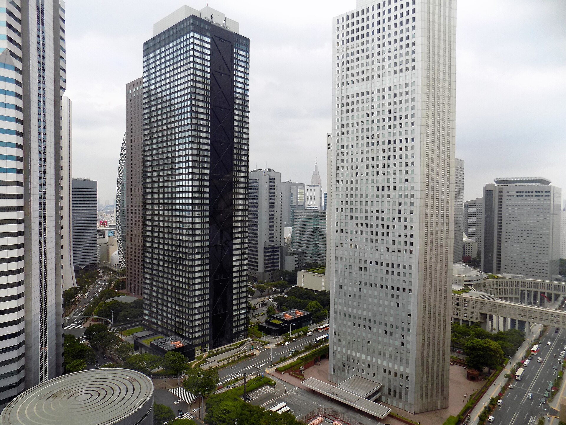 The Nishi-Shinjuku skyscraper cluster seen from above, office towers and elevated walkways under a pale sky